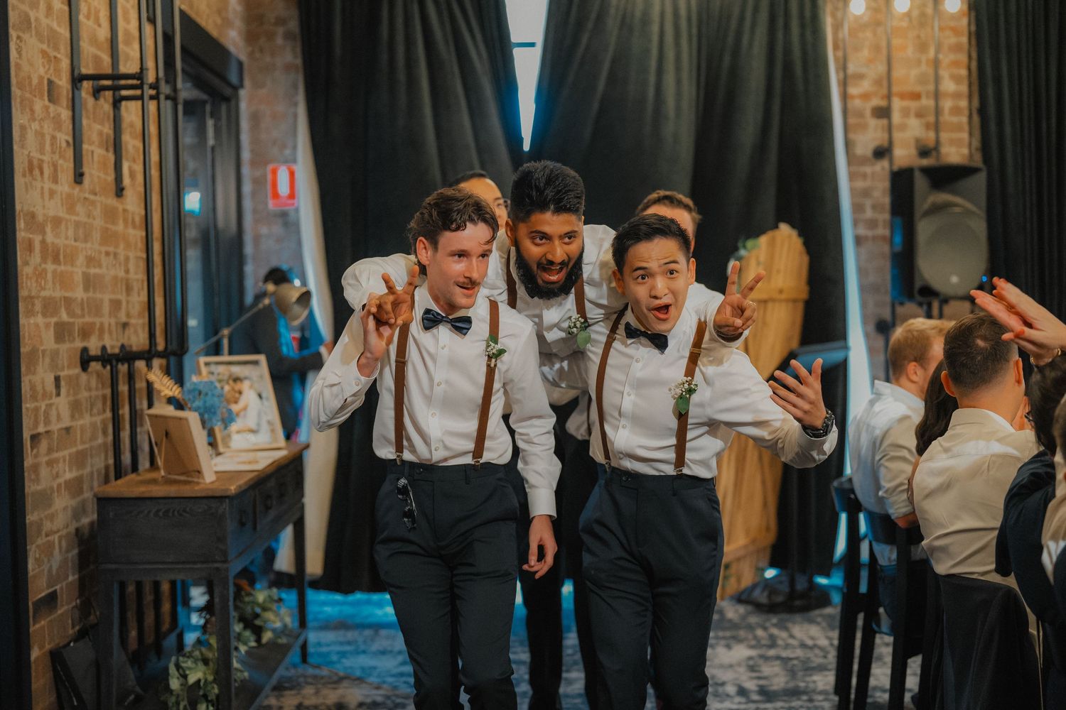 Groomsmen in suspenders and bowties walking through an industrial-style space with brick walls.