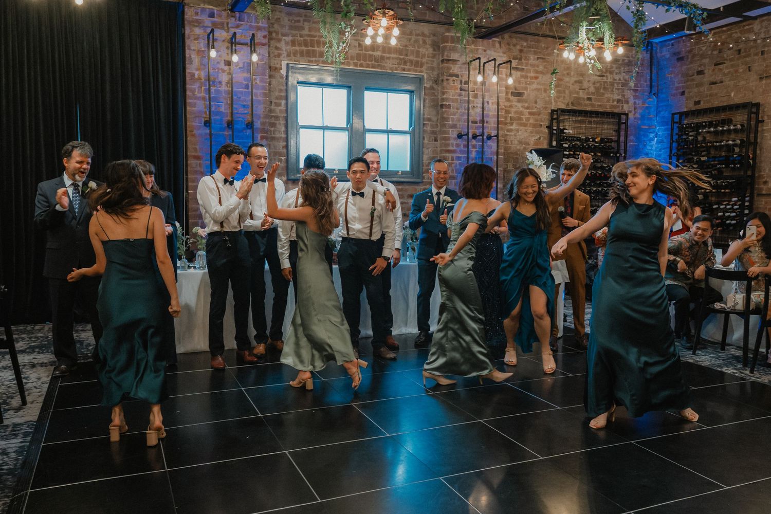 Wedding guests dancing together on a dark dance floor in an industrial-style venue with exposed brick walls.