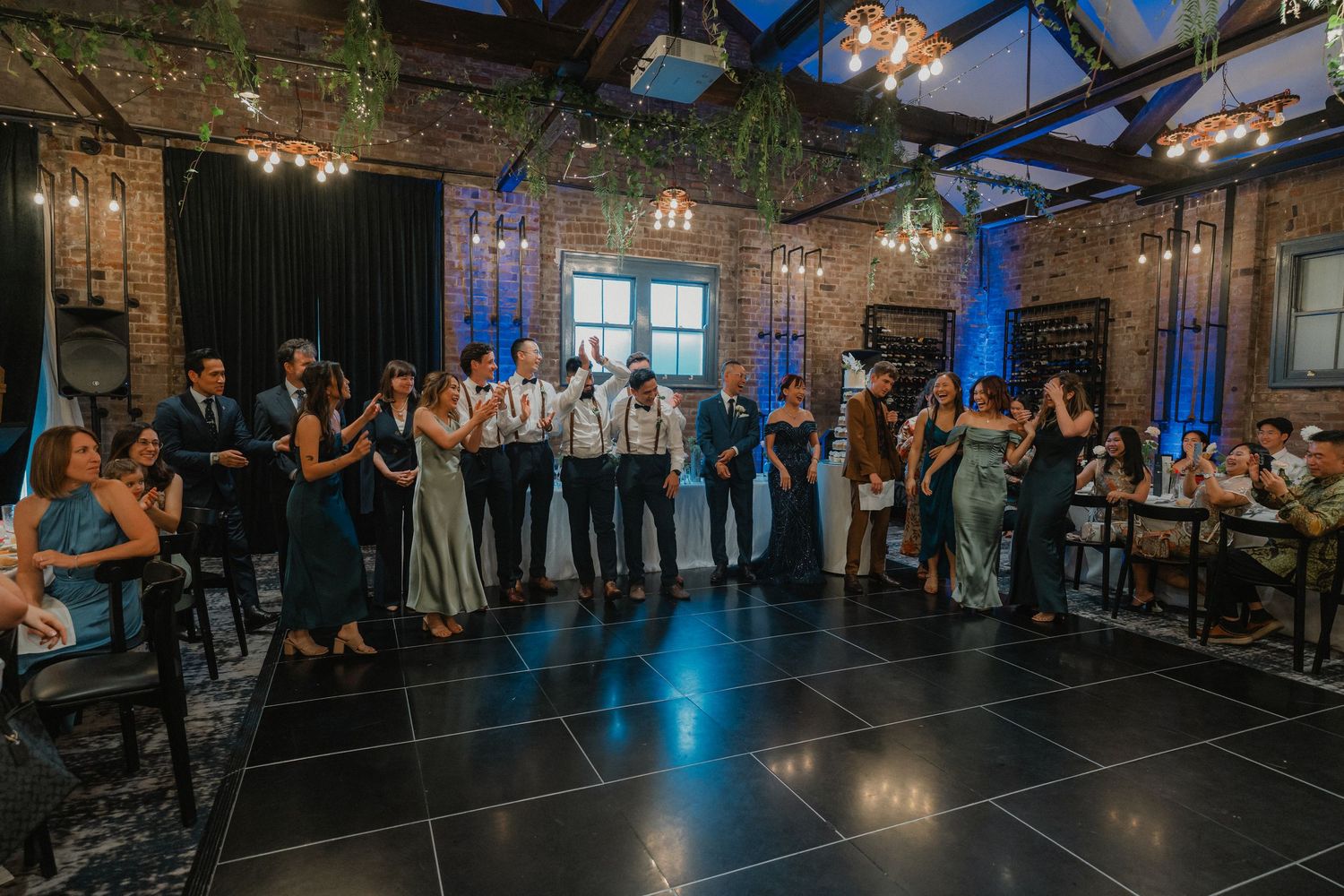 Wedding guests gather around an illuminated dance floor in an industrial-style venue with hanging lights and greenery.
