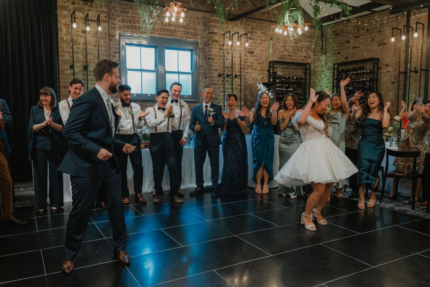 Couple dances at wedding reception in industrial brick venue with chandeliers and guests watching in background.