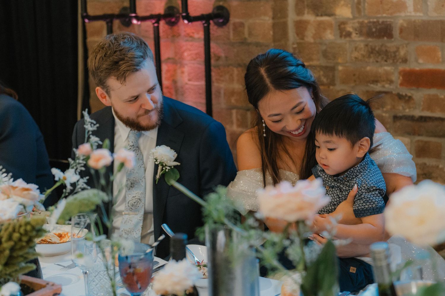 Intimate moment at wedding reception dinner with floral centerpieces and exposed brick wall backdrop.