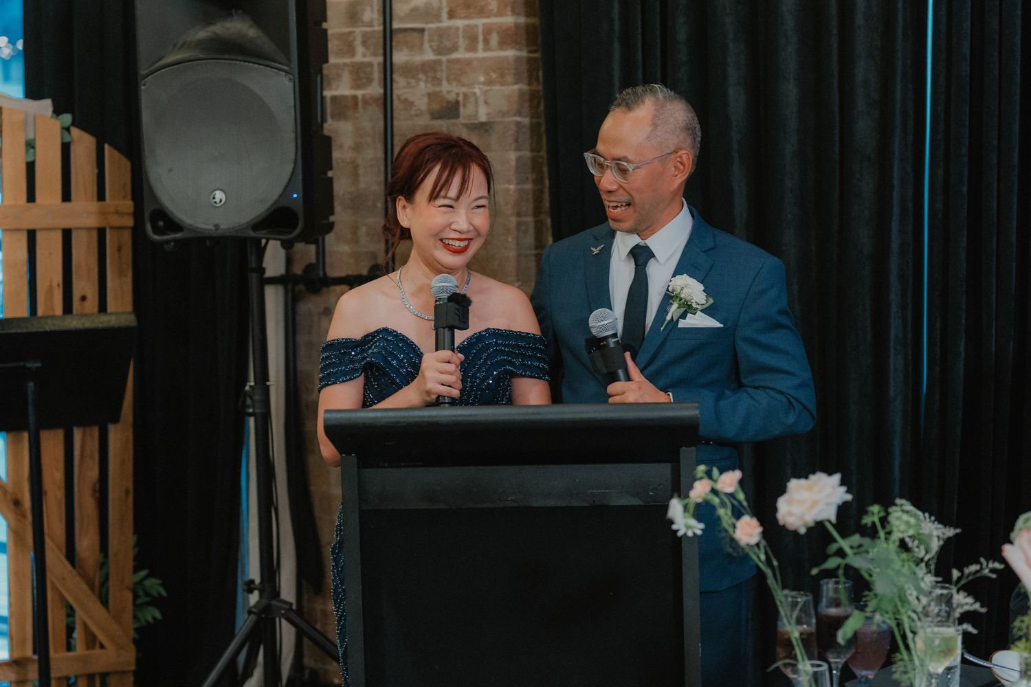 Speakers at wedding podium with white flowers and navy blue formal attire against exposed brick wall.