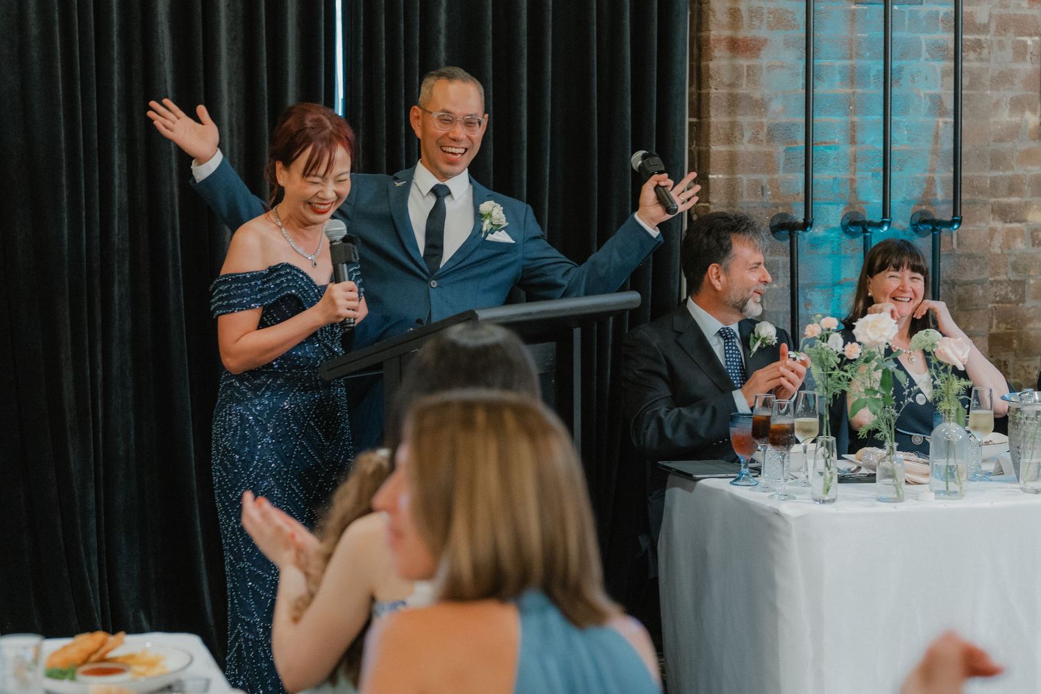 Wedding reception speech with guests seated at decorated tables in industrial-style venue.