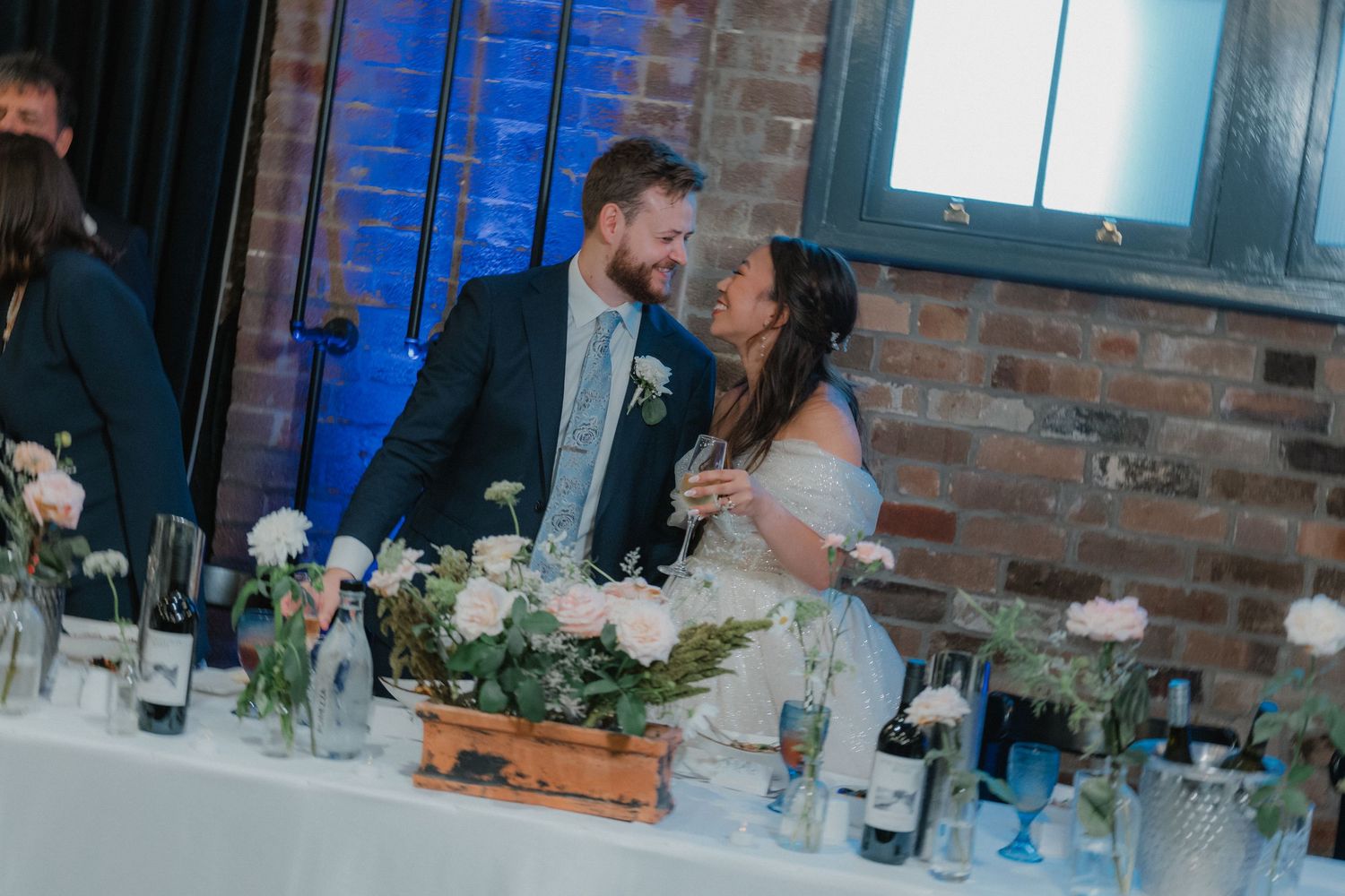 Joyful moment at wedding reception head table with floral centerpieces and exposed brick backdrop.