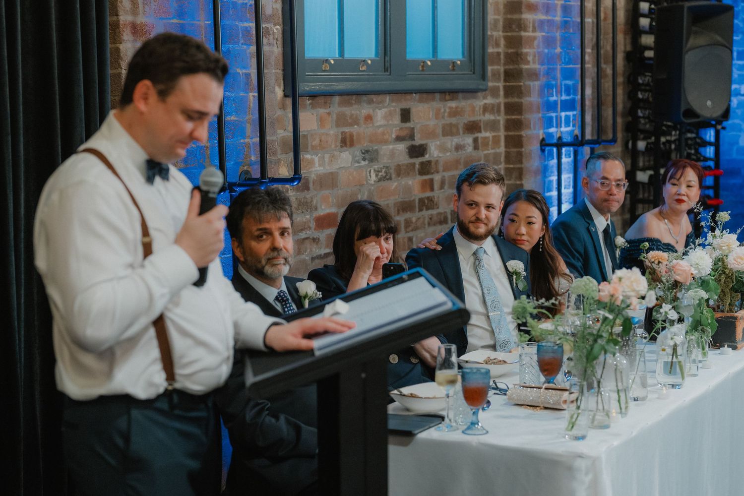 Guest delivers speech at wedding reception with brick wall backdrop and floral table decorations.