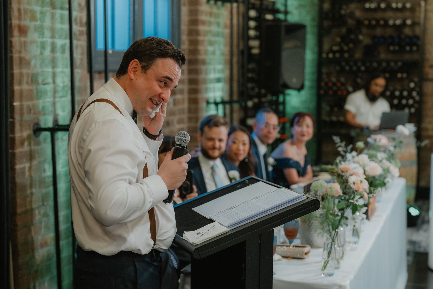 A wedding reception speech being delivered at an industrial-style venue with exposed brick walls.