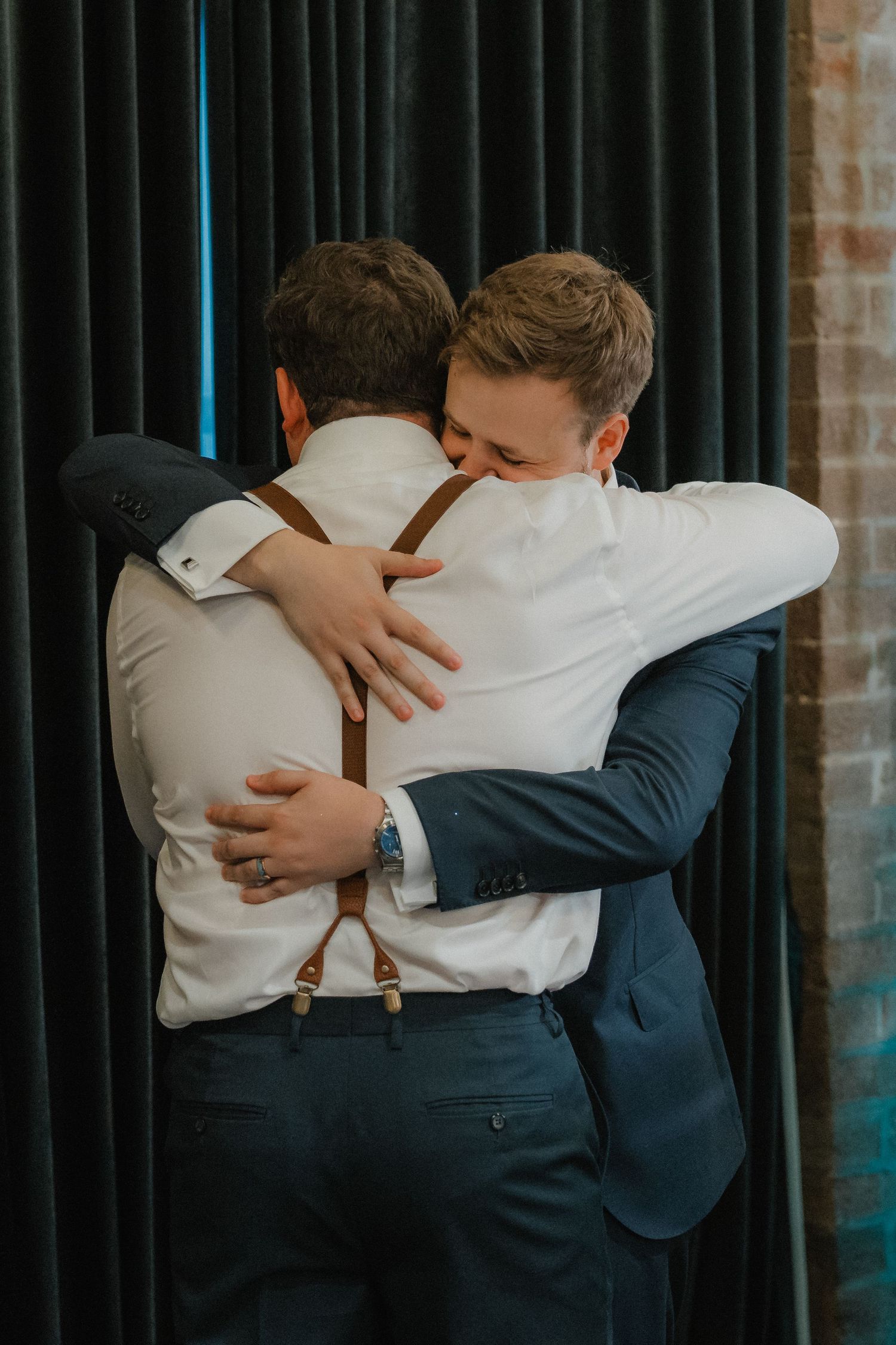 Emotional embrace between two people in formal attire against black curtain backdrop.