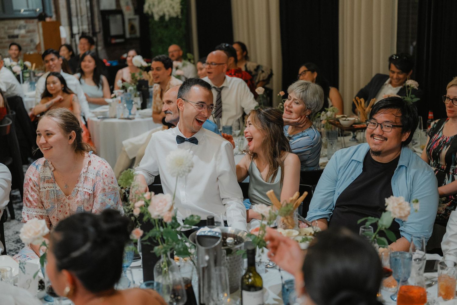 Wedding guests enjoying a reception dinner at tables decorated with greenery in an elegant venue.
