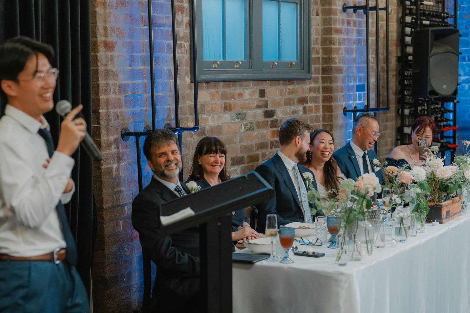 Wedding reception with toast being given while guests seated at decorated tables in brick-walled venue.