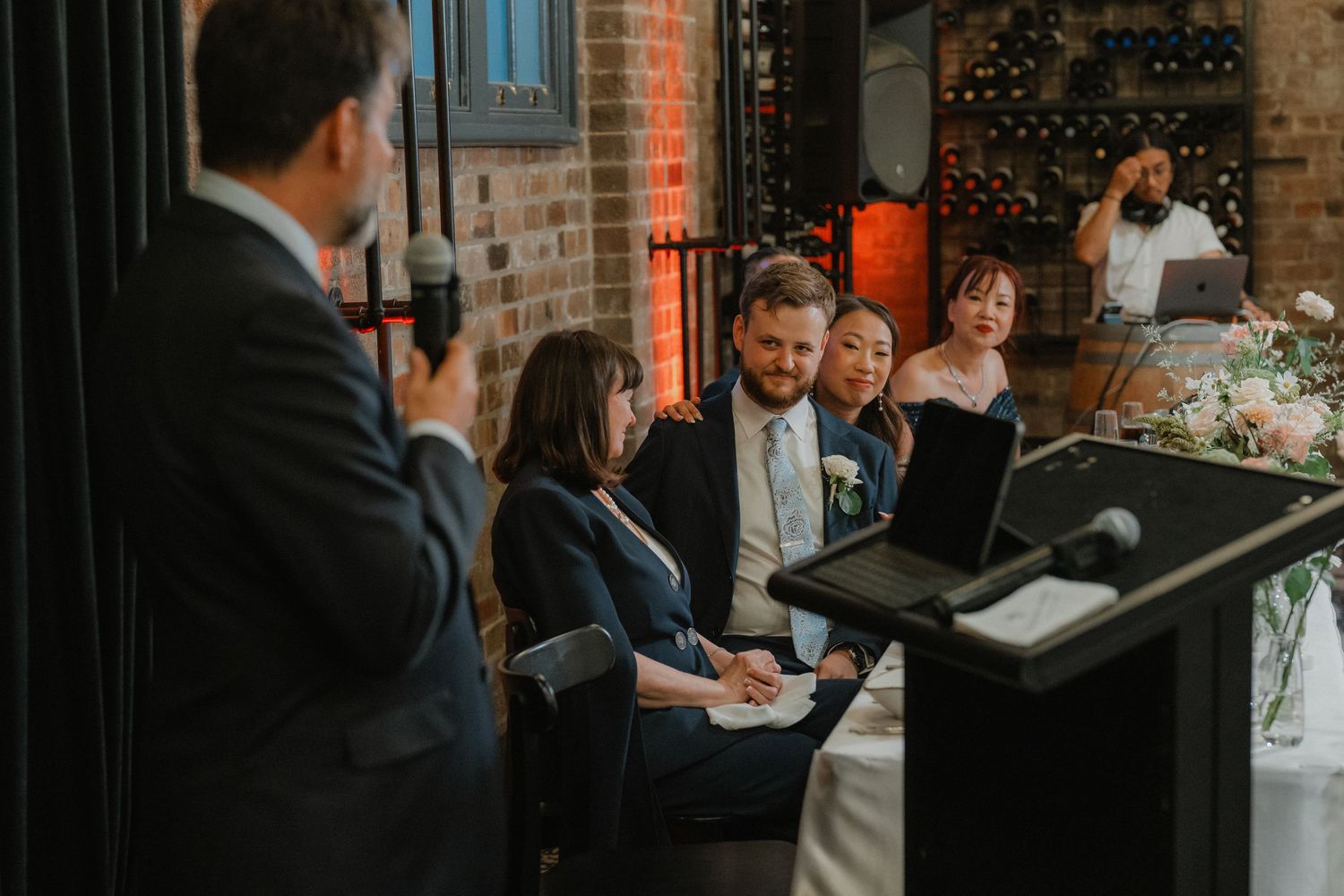 Wedding reception speech in brick-walled venue with ambient lighting and floral decorations.