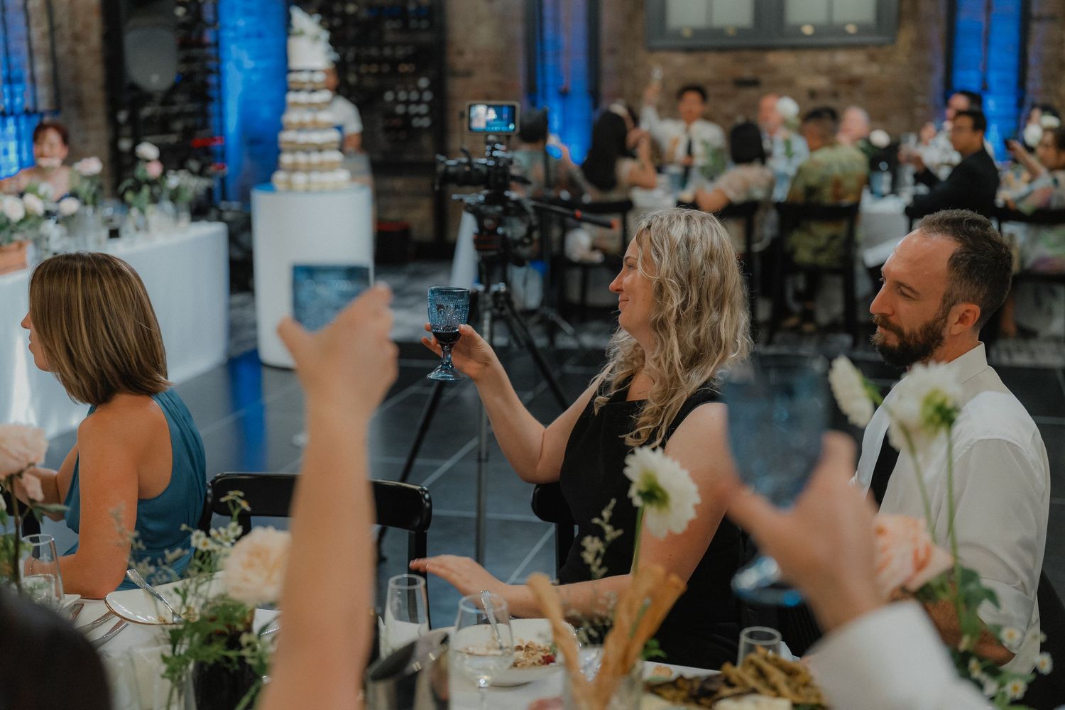 Wedding reception guests raising glasses for a toast in an elegant blue-lit venue.