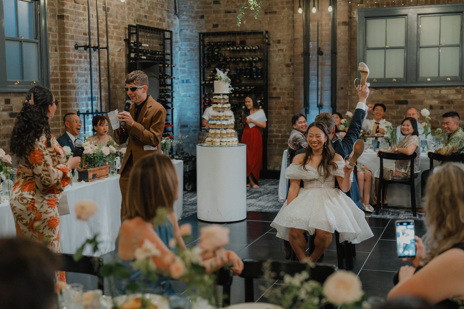 Wedding reception in industrial venue with exposed brick walls and tiered white cake display.