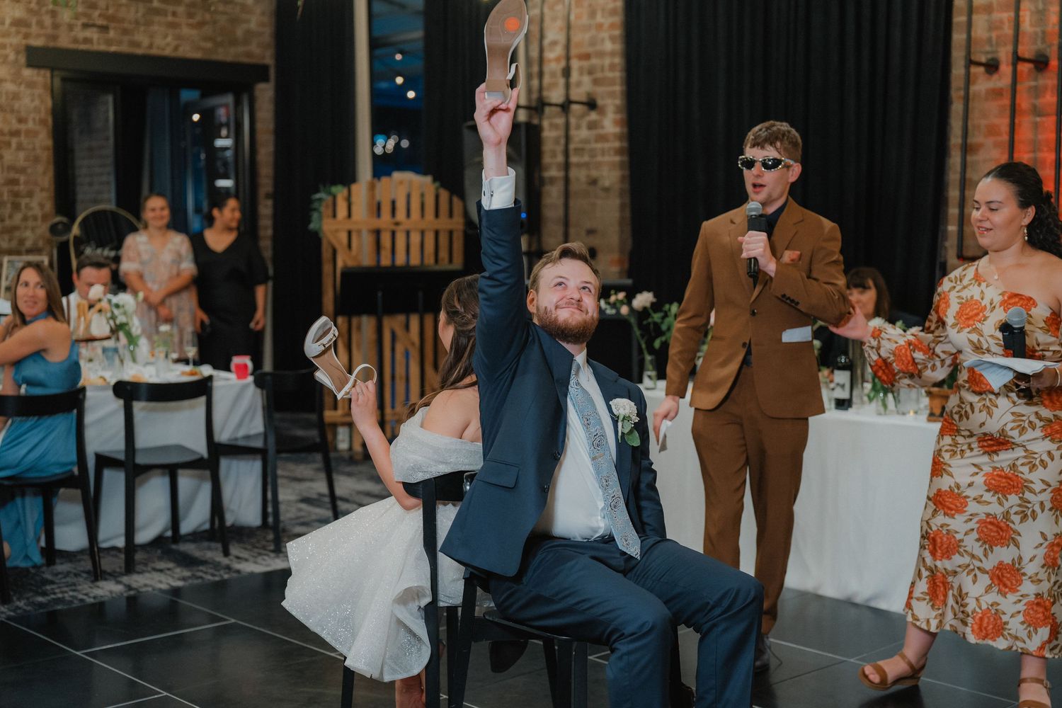 Wedding reception guests mingling at tables in an industrial-style venue with brick walls.
