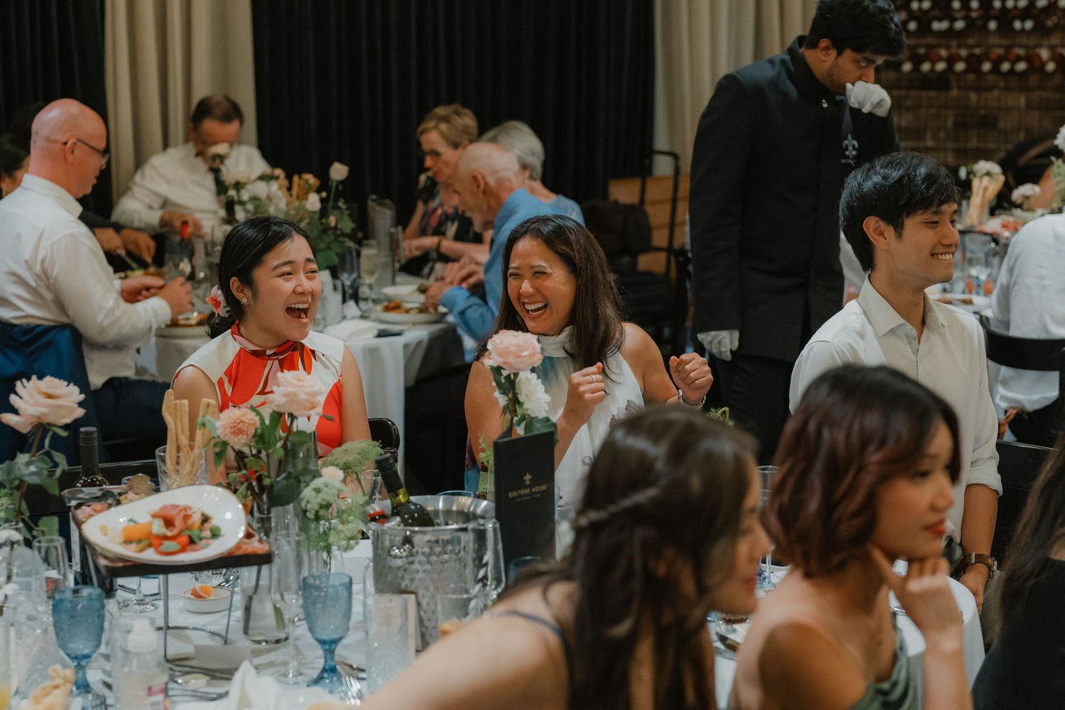 Wedding reception guests enjoying dinner and conversation at elegantly decorated tables in a dimly lit venue.
