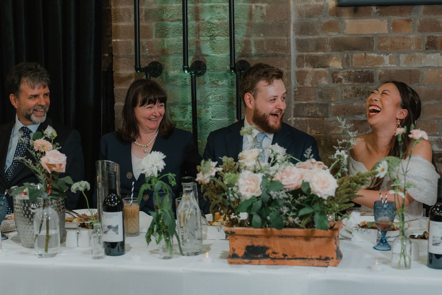 Wedding guests share a candid moment of laughter at the reception dinner table decorated with floral centerpieces.