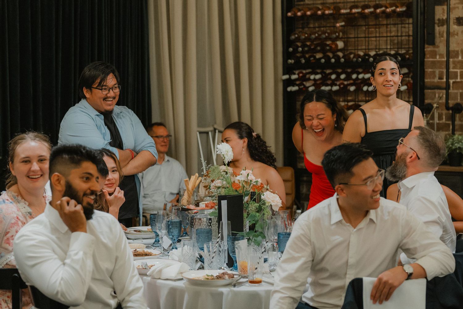 Wedding guests seated at elegant tables share laughter and conversation during a reception dinner.