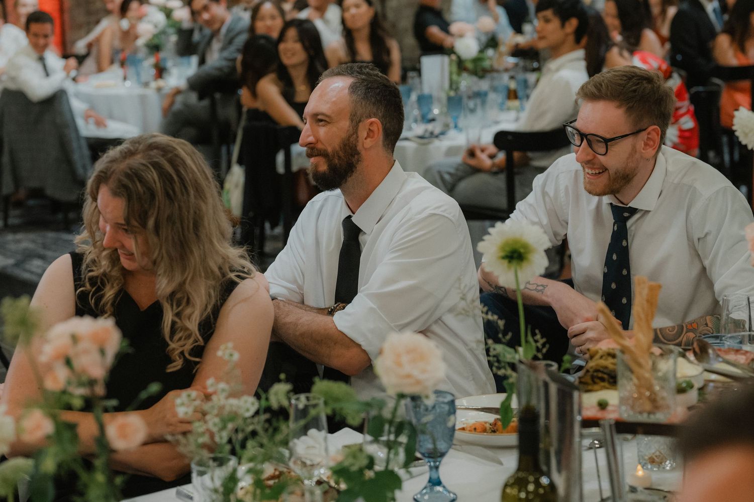 Wedding reception guests laugh and smile during dinner at elegantly decorated tables with white floral centerpieces.
