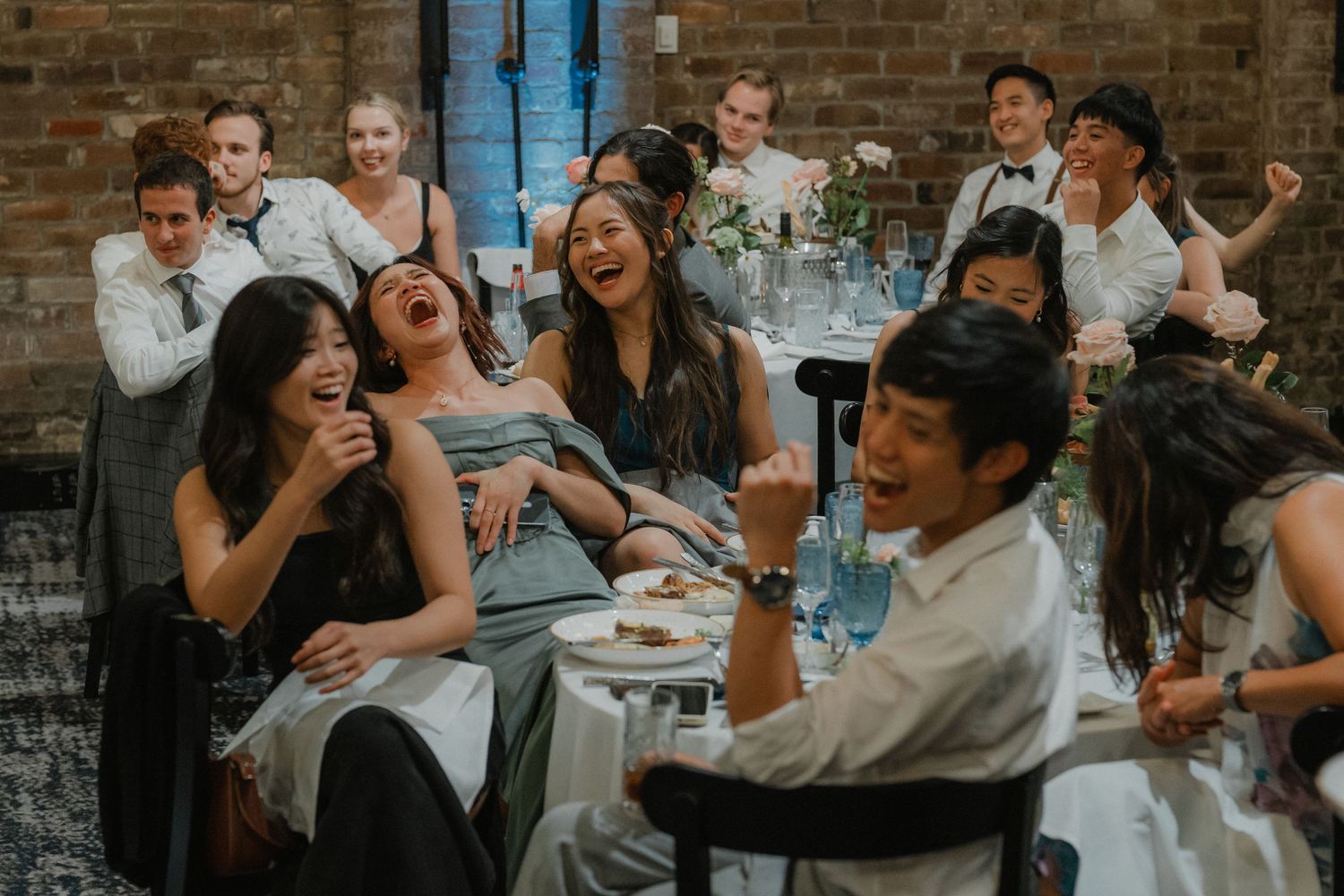 Guests laugh and celebrate during wedding speeches at long tables in an industrial-style venue.