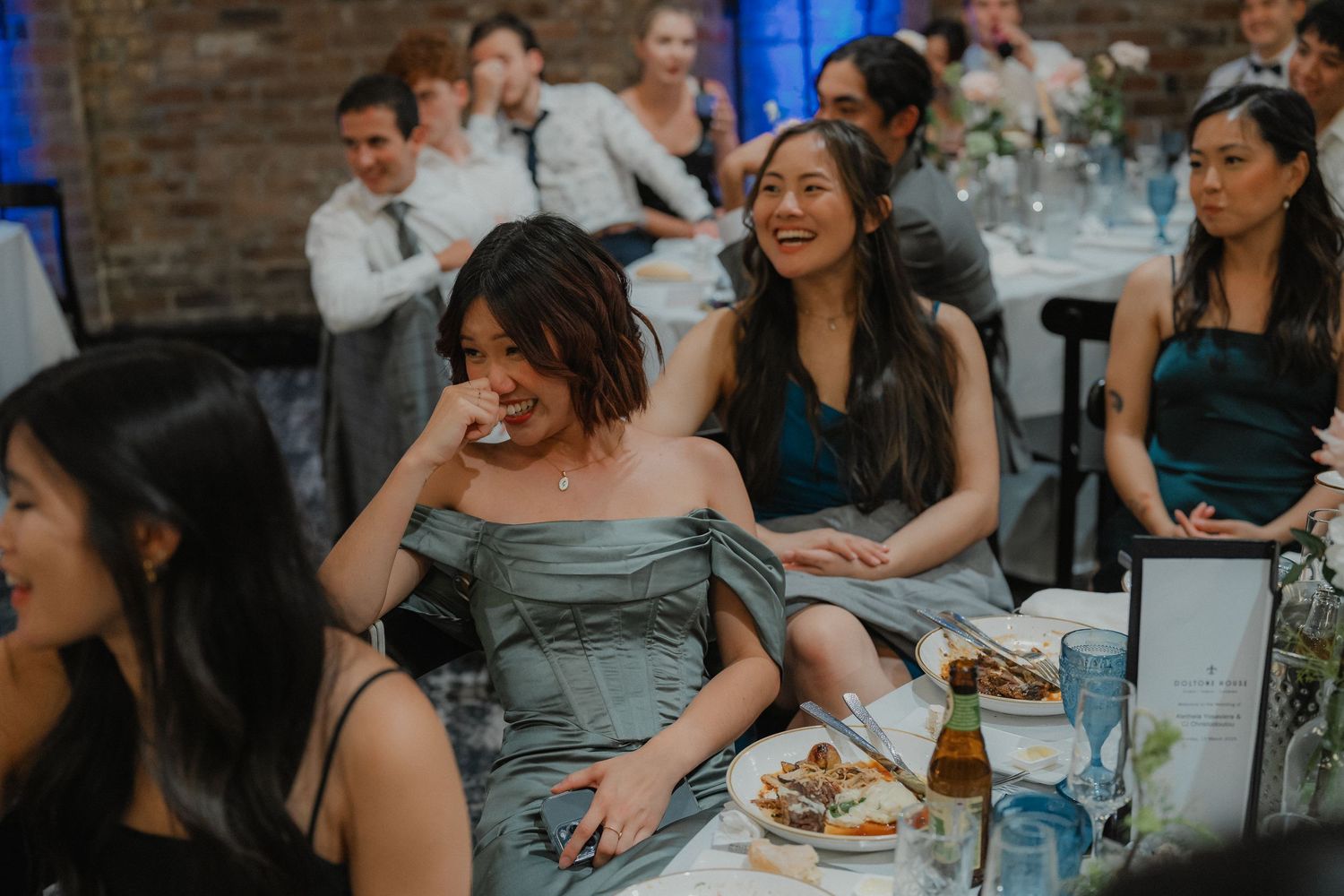 Wedding guests share laughter and joy at a reception table with brick walls and blue decor.