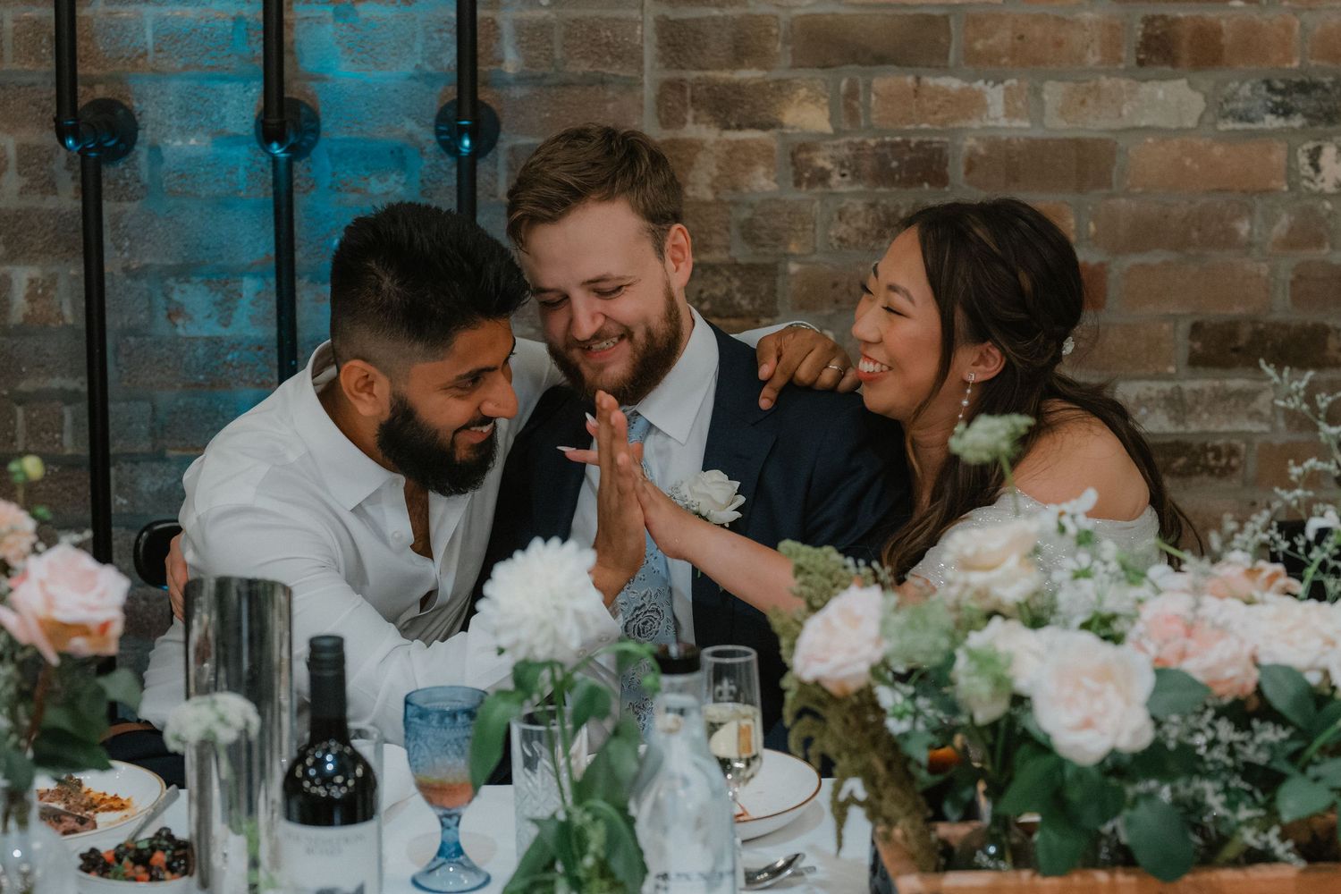 Wedding guests share joyful moments at tables decorated with white floral arrangements against exposed brick walls.