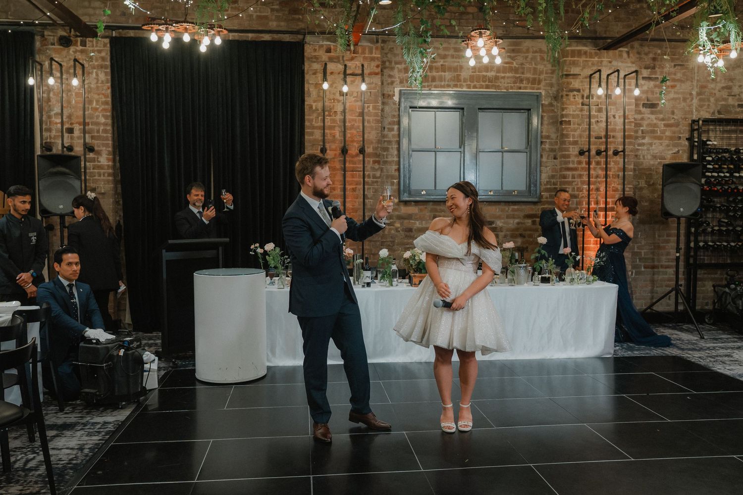 A couple performs their first dance in an industrial-style venue with brick walls and string lights.