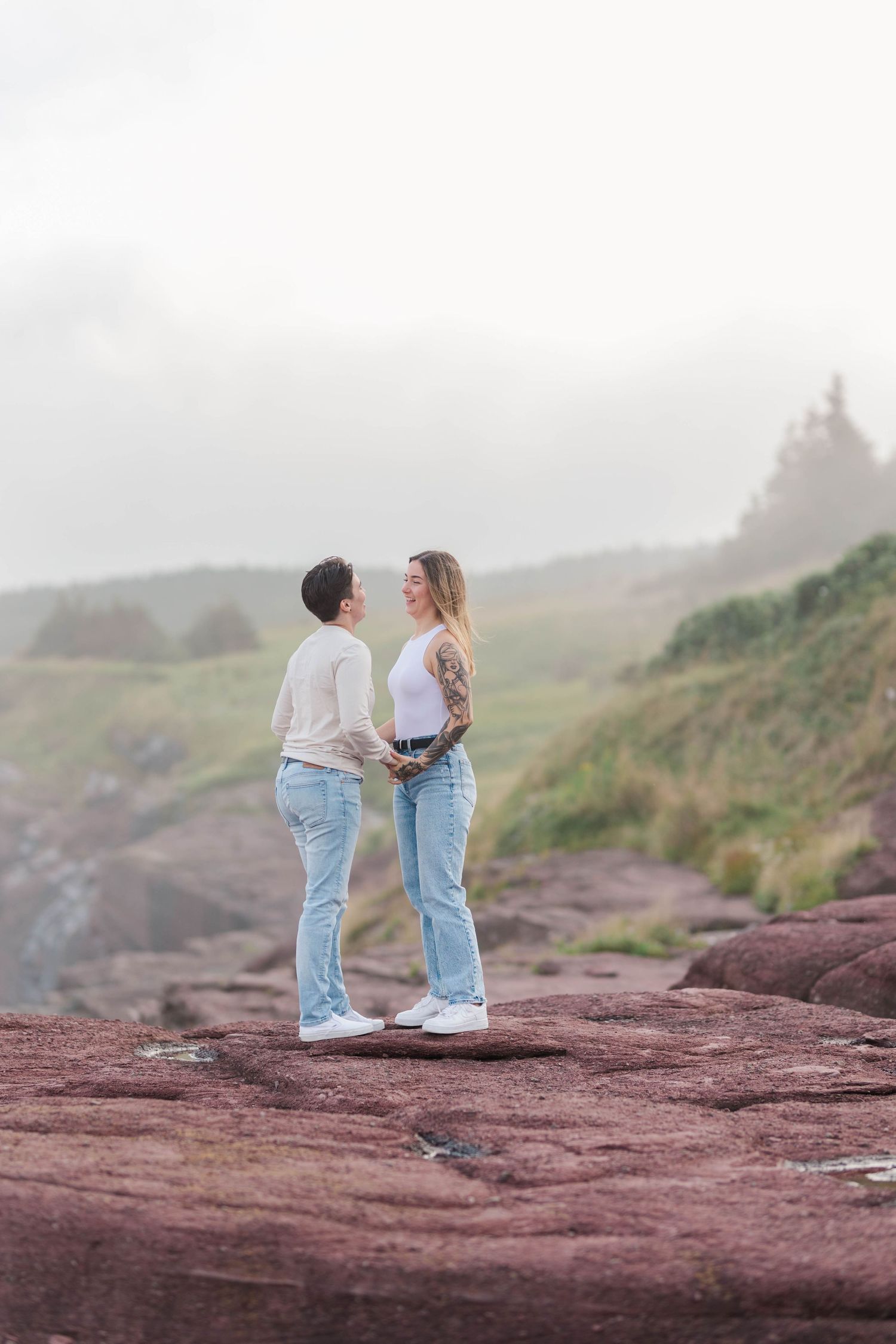 Couple standing together on red rocky cliffs of Flatrock, NL during a misty coastal evening.