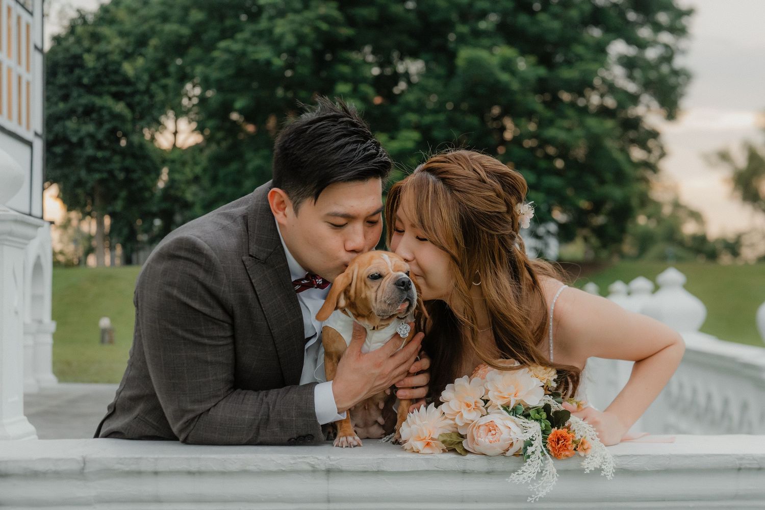 A couple in wedding attire cuddles with their small brown dog on marble steps outdoors during sunset.