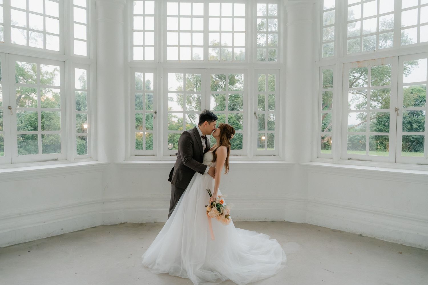 A romantic wedding moment captured in a bright sunlit conservatory with white windows and elegant architectural details.