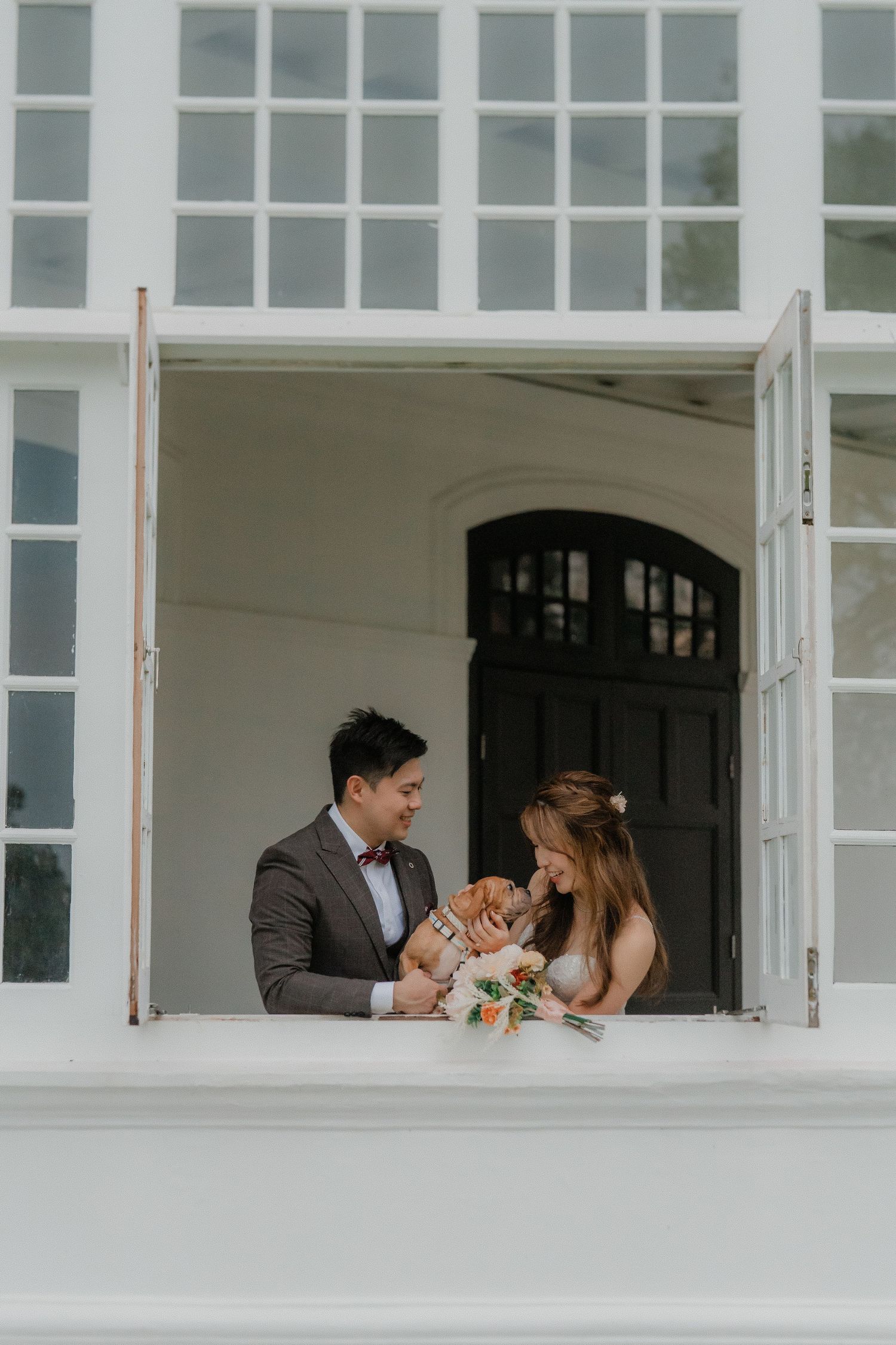 A wedding couple and their dog share an intimate moment at a window archway of a white building.