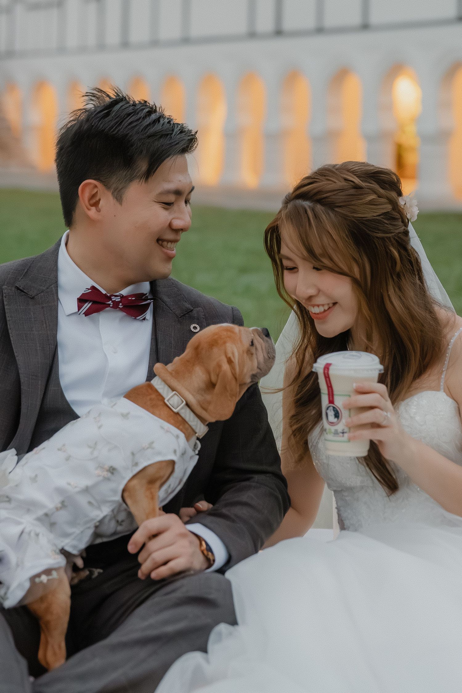 A happy couple in wedding attire shares laughter and coffee outdoors during their special day.