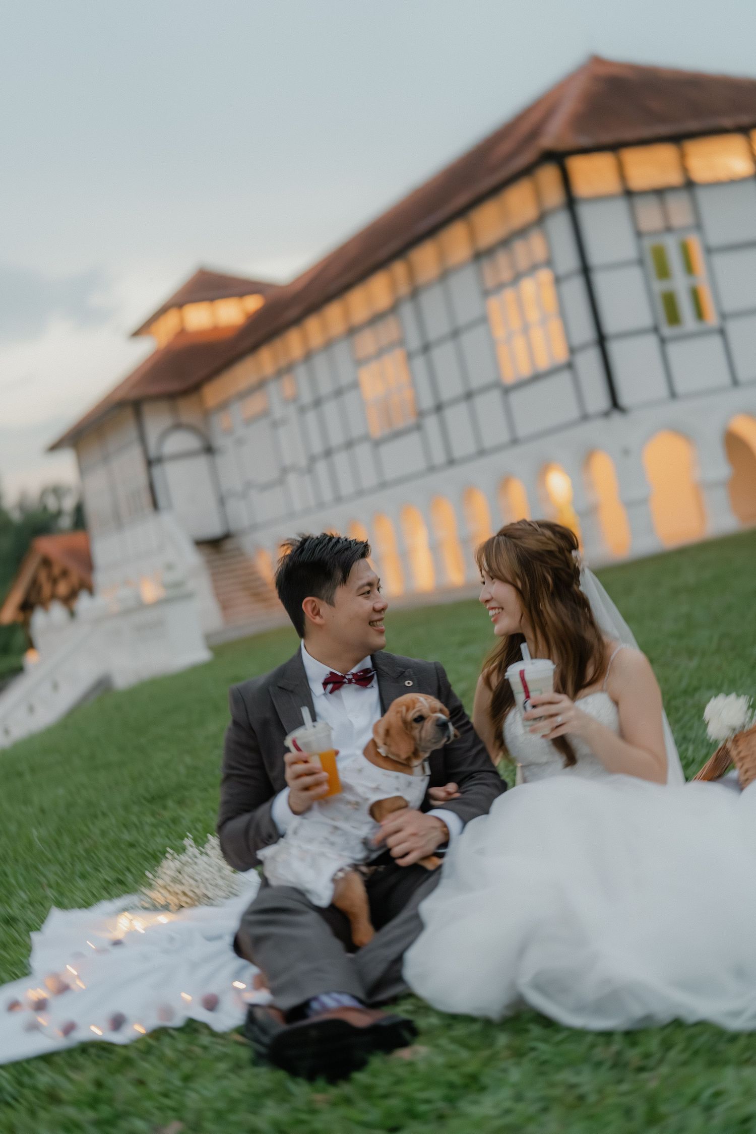 Wedding couple relaxes on grass with their corgi while having drinks near white building.