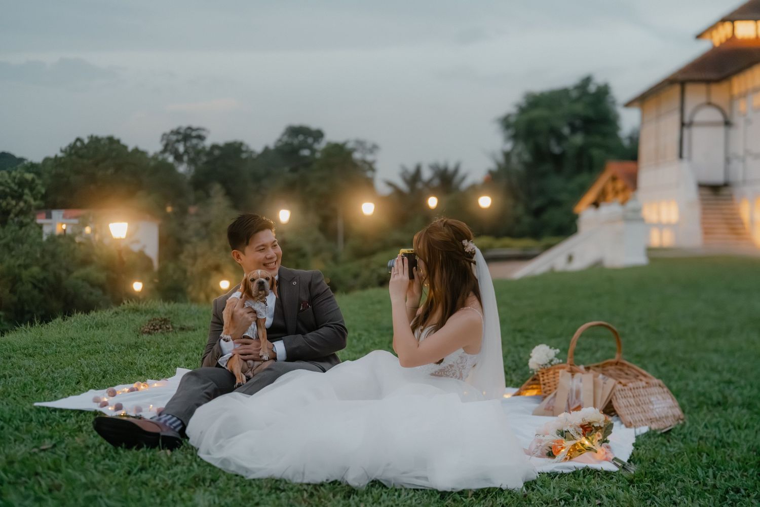 Wedding couple enjoys picnic on lawn at dusk with illuminated building backdrop.