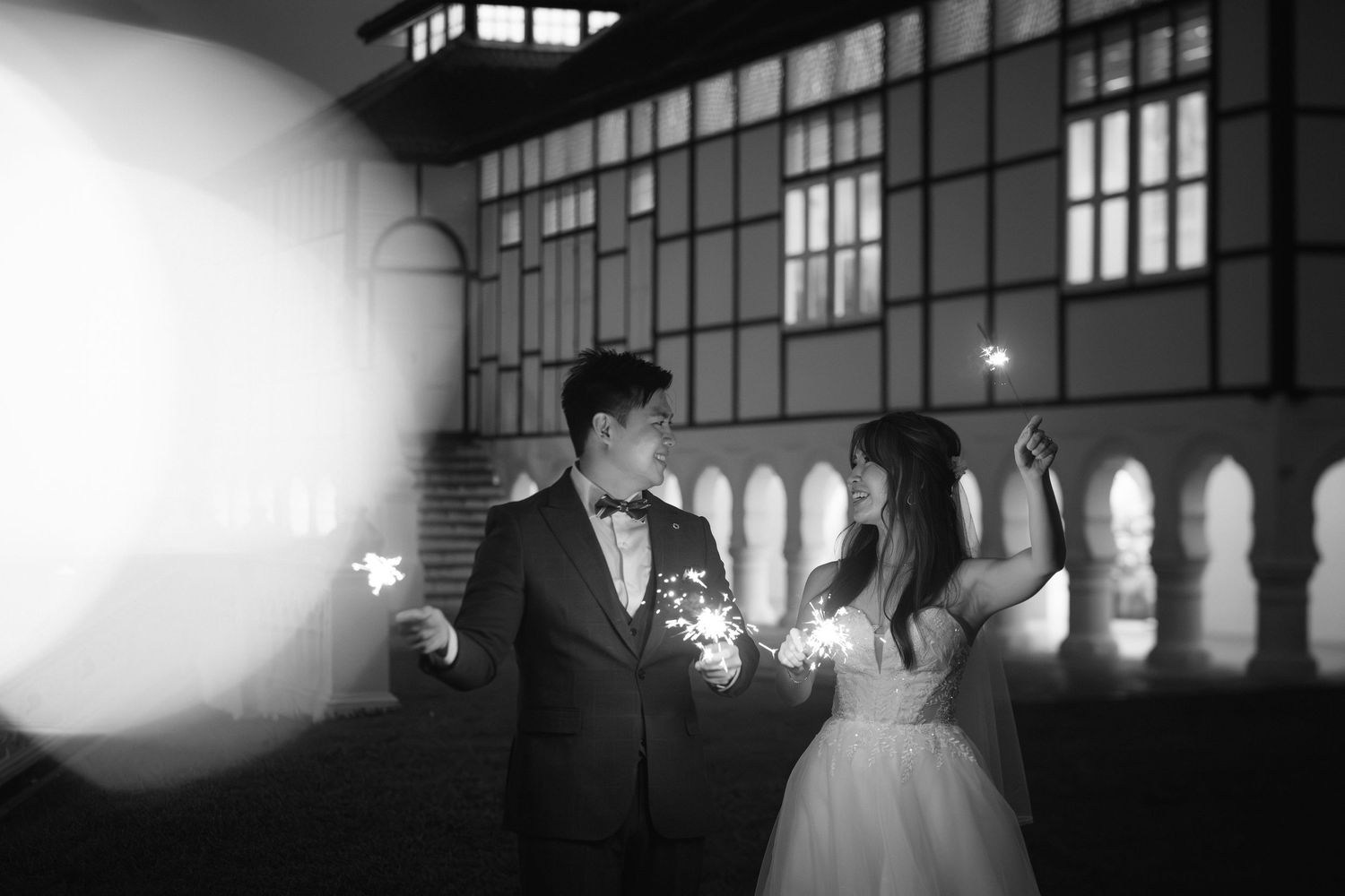 A couple in formal wedding attire hold sparklers while standing in an elegant venue with arched windows at night.
