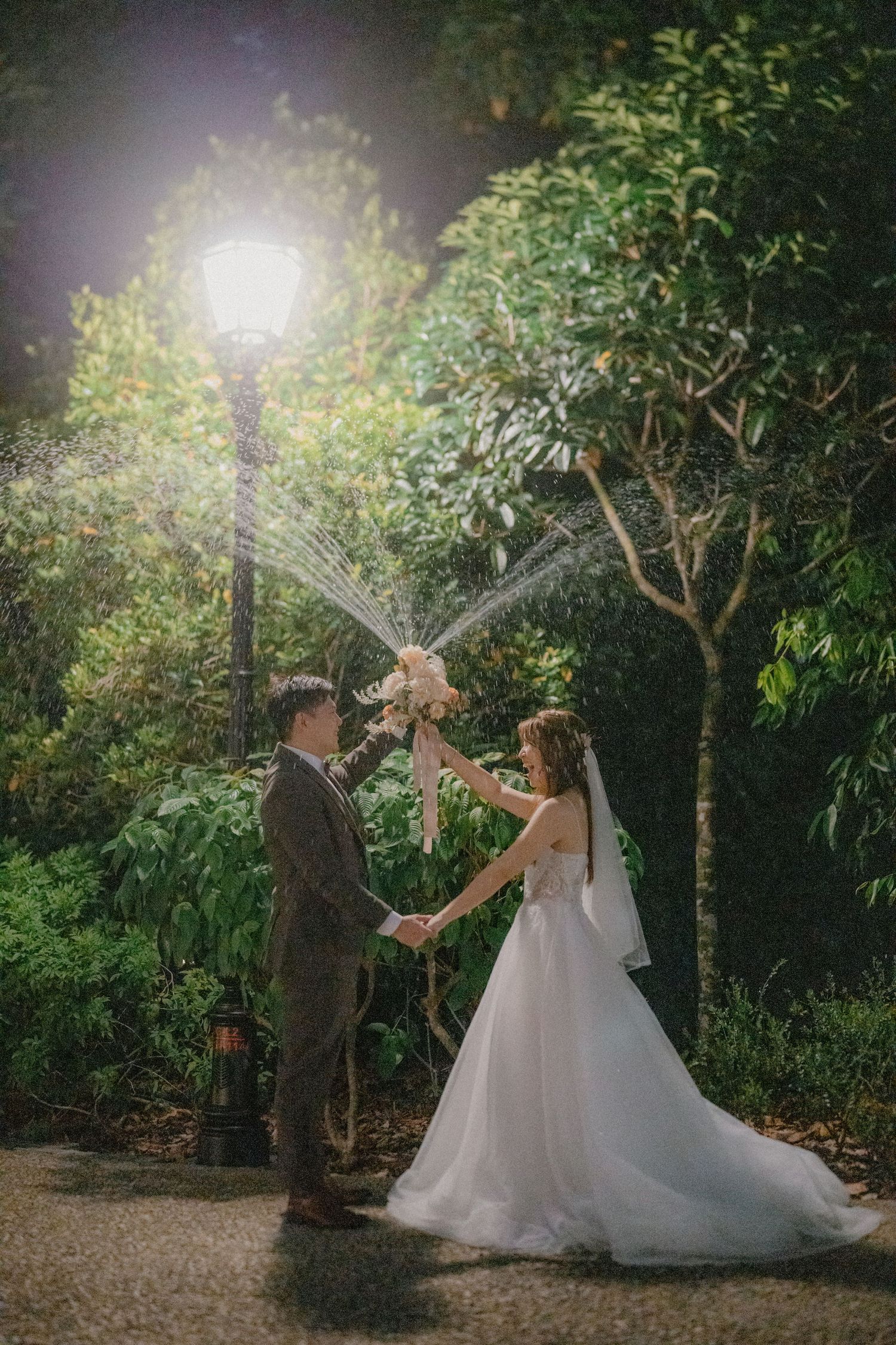 A romantic wedding couple stands beneath a glowing lamppost in a lush garden setting at night.
