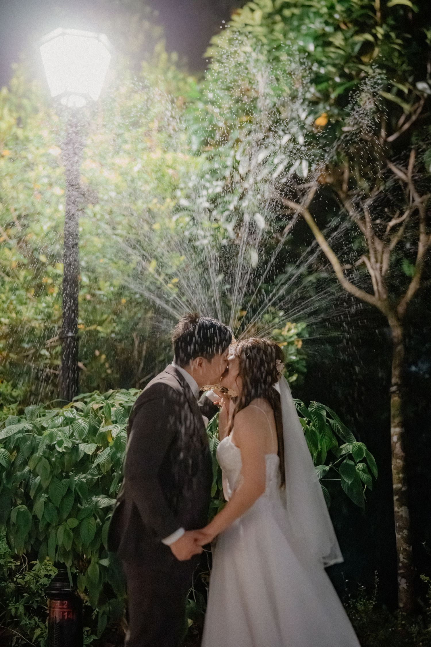 A couple shares a romantic moment under garden sprinklers during their nighttime wedding photoshoot.