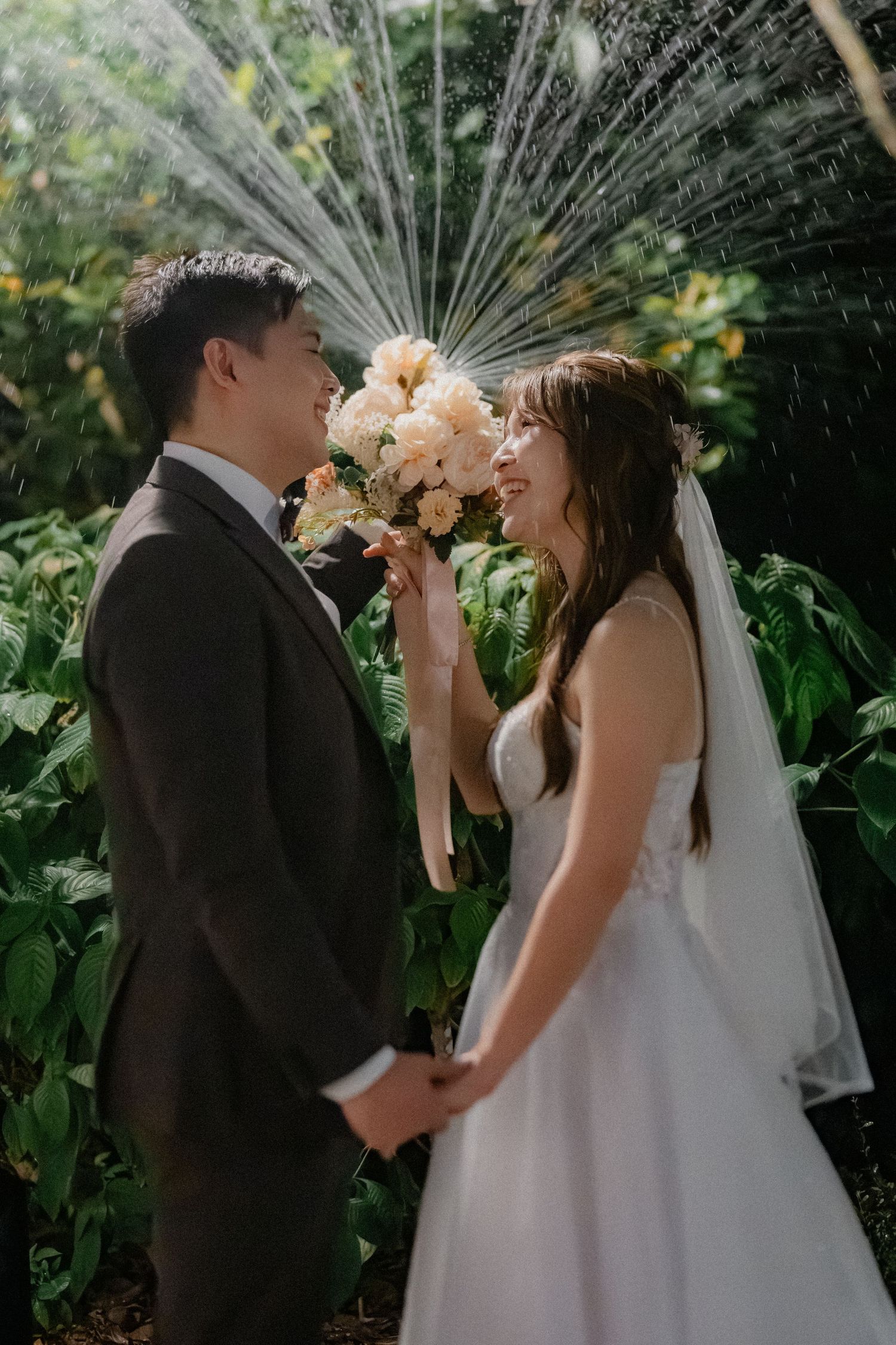 Wedding couple posing in a garden with sprinklers creating a misty backdrop of water droplets.