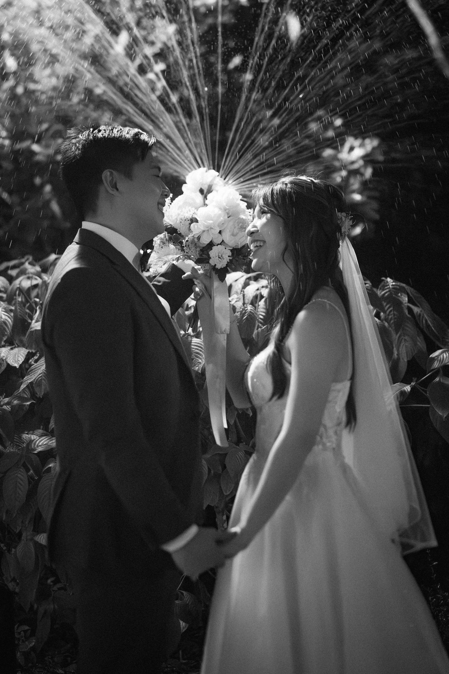 A romantic black and white wedding photo showing a couple holding hands with a floral bouquet under sunlit trees.