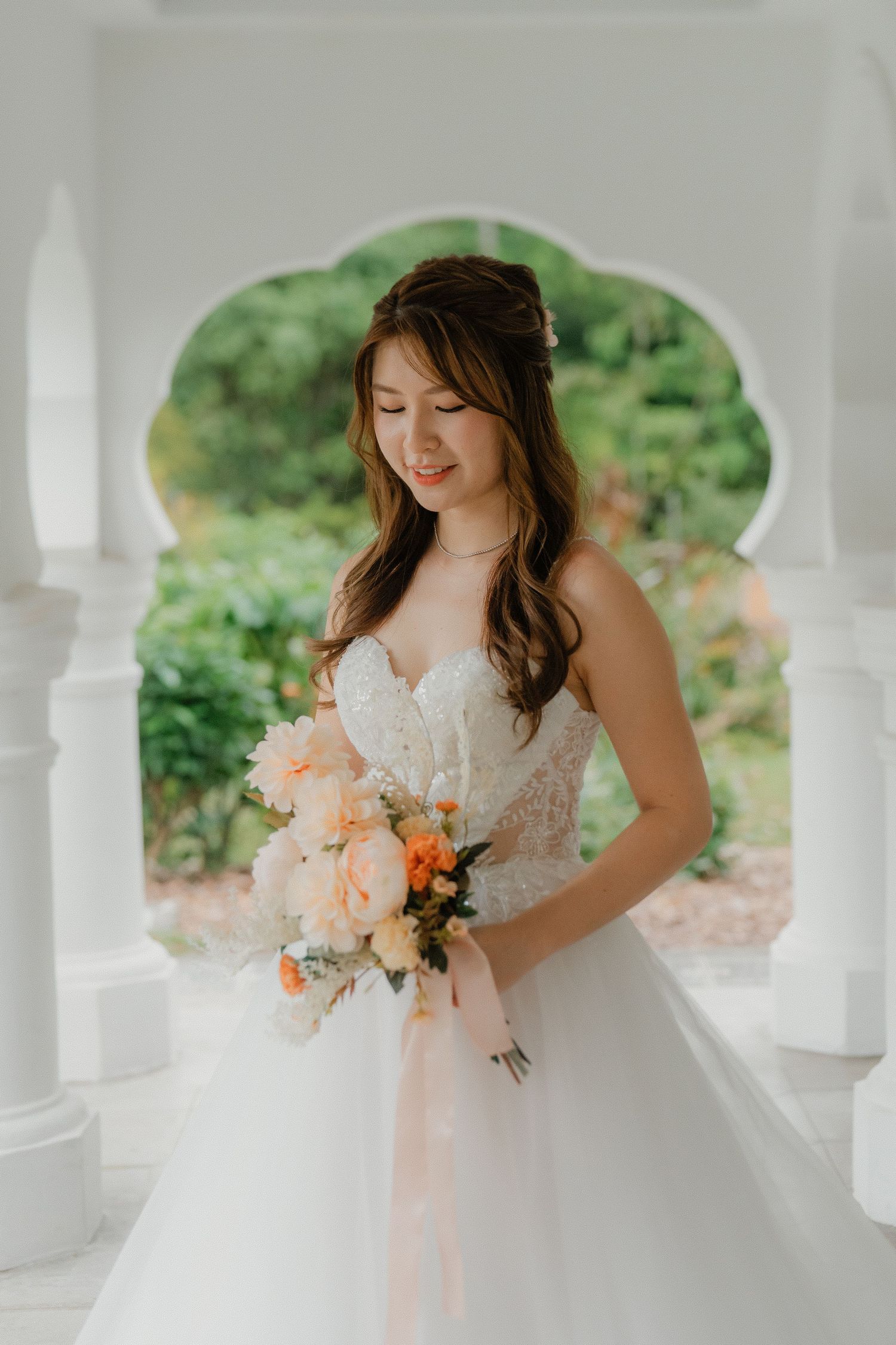 A bride in a flowing white wedding gown holds a peach and cream bouquet while standing on an elegant white porch.