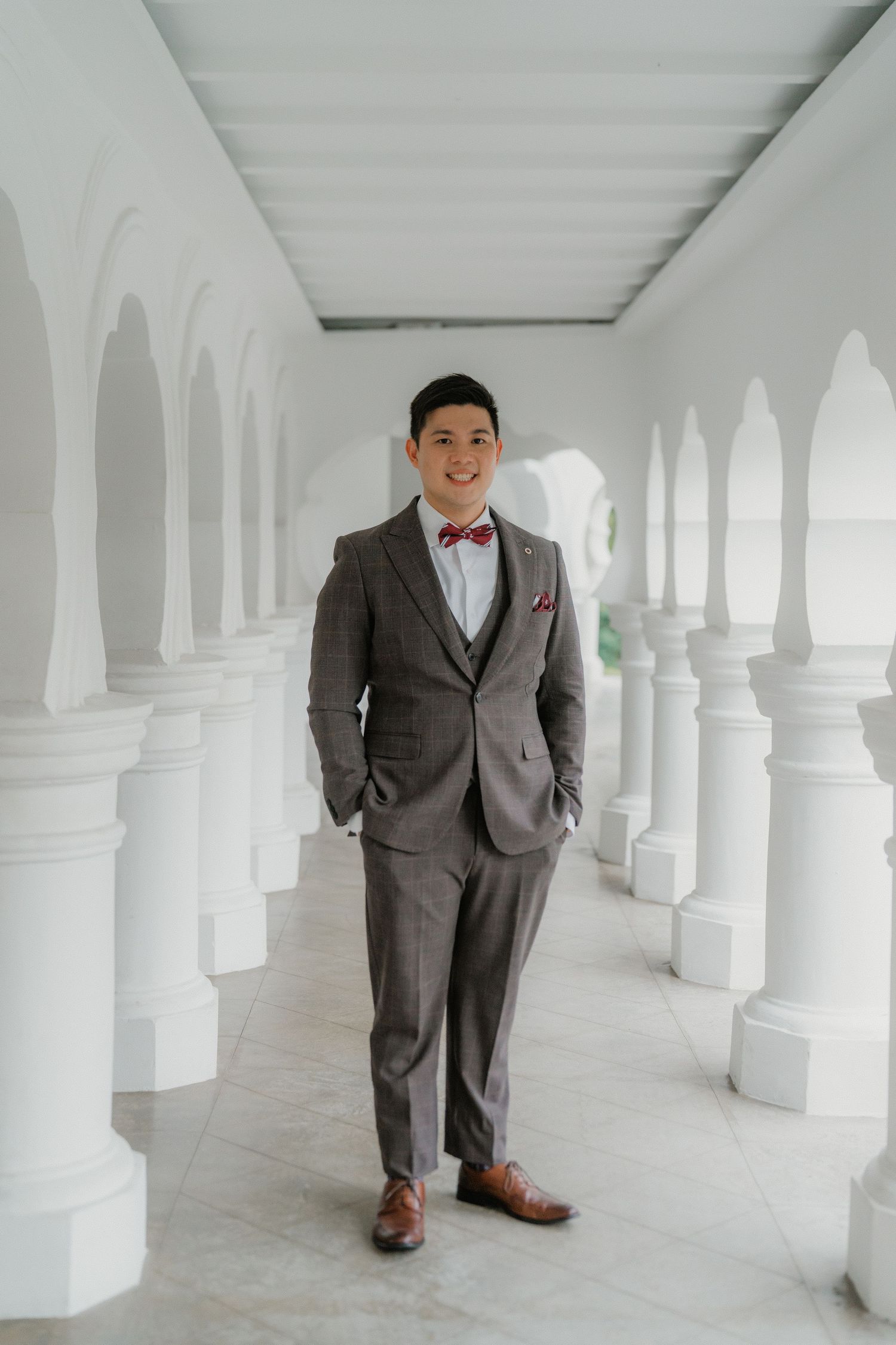 Groom in a grey suit with red bowtie poses in a white columned hallway.