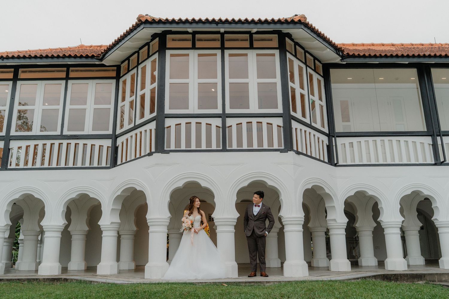 An elegant couple stands beneath a white arched colonnade of a Spanish colonial style building with a tiled roof.