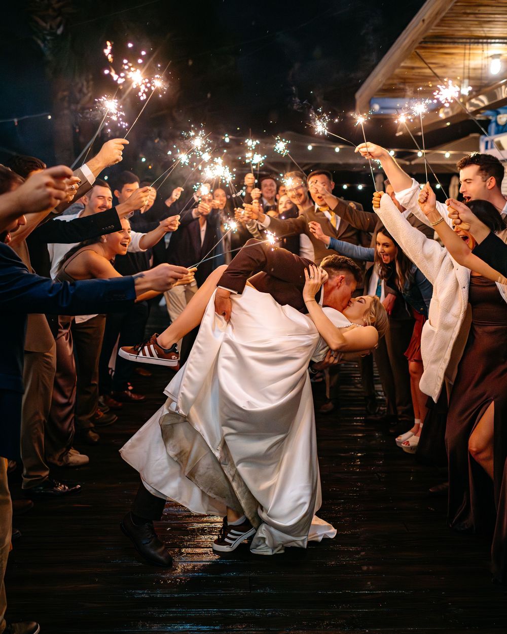 Bride and groom share a romantic dip kiss during a sparkler send-off surrounded by cheering wedding guests at night.