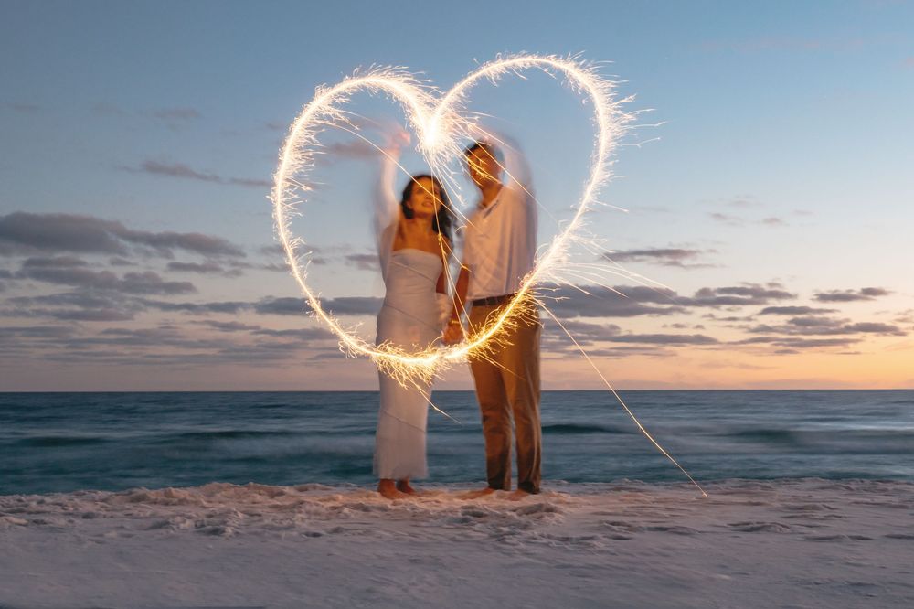 Couple creates heart shape with sparklers on beach during sunset while waves crash in background.