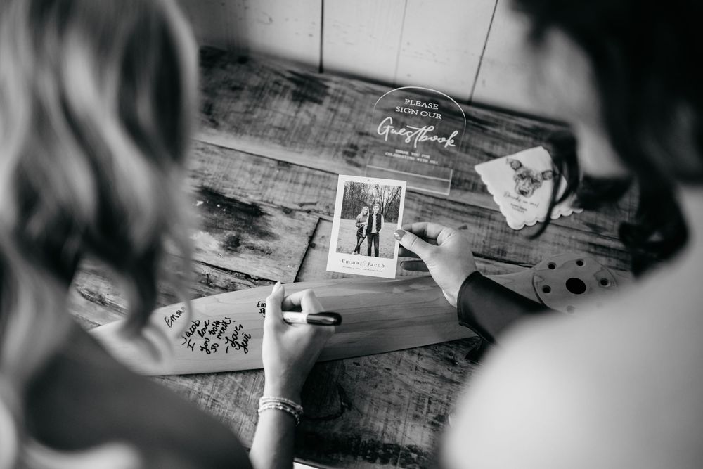Black and white photo of a bride signing a wedding guest book with Polaroid photos on a rustic wooden table.