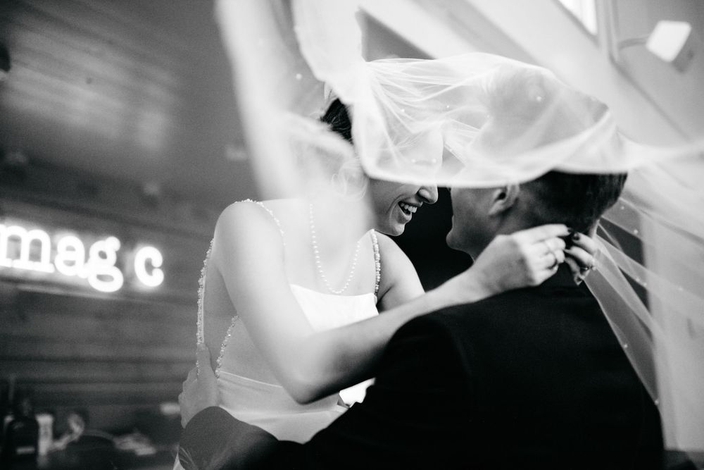 Black and white photo of bride and groom sharing a kiss under a wedding veil at a romantic venue.