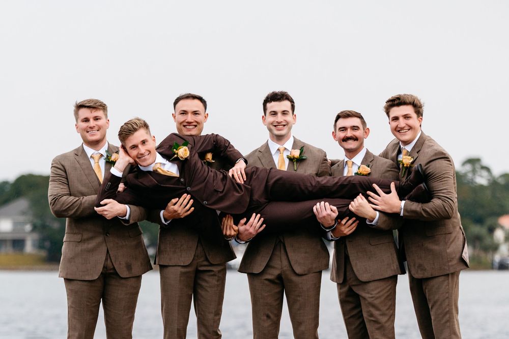 Groomsmen in brown suits lifting the groom outdoors near a waterfront on a wedding day.
