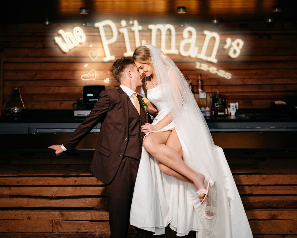 Newlyweds share a romantic kiss under a glowing neon sign reading The Pitmans at their wedding venue.