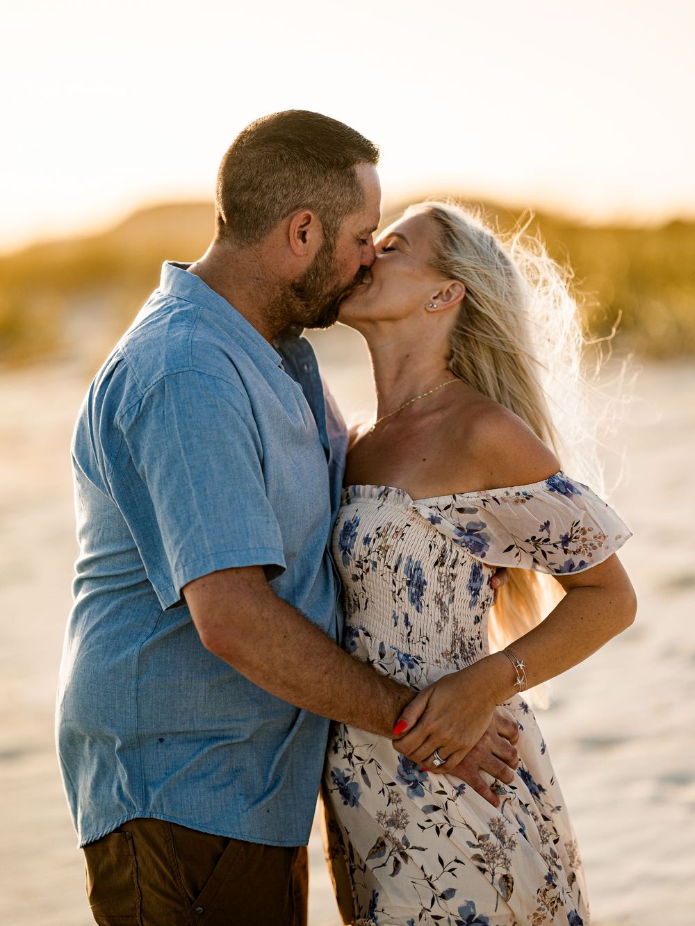 Couple sharing a romantic kiss at sunset on the beach, woman in floral off-shoulder dress, golden hour lighting.