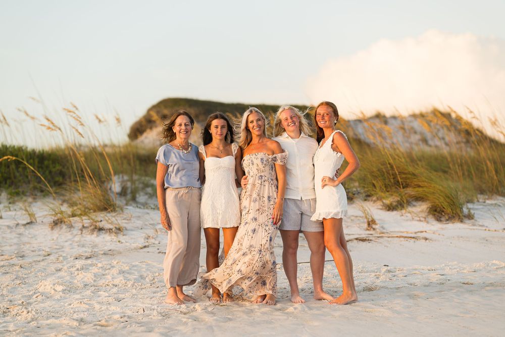 Five women posing together on a sandy beach at golden hour near sea oats and white sand dunes.