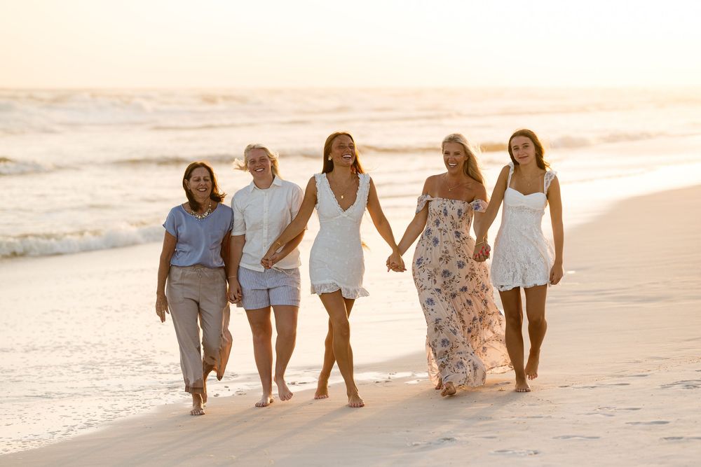 Five women walking together on a sunny beach at sunset, wearing white and casual outfits near ocean waves.