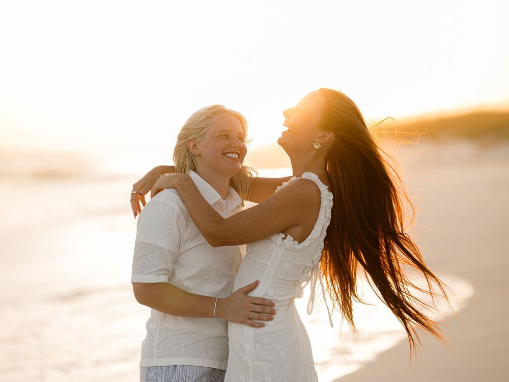 Two women laughing and embracing on a sunny beach at golden hour, wearing white outfits with ocean waves behind them.